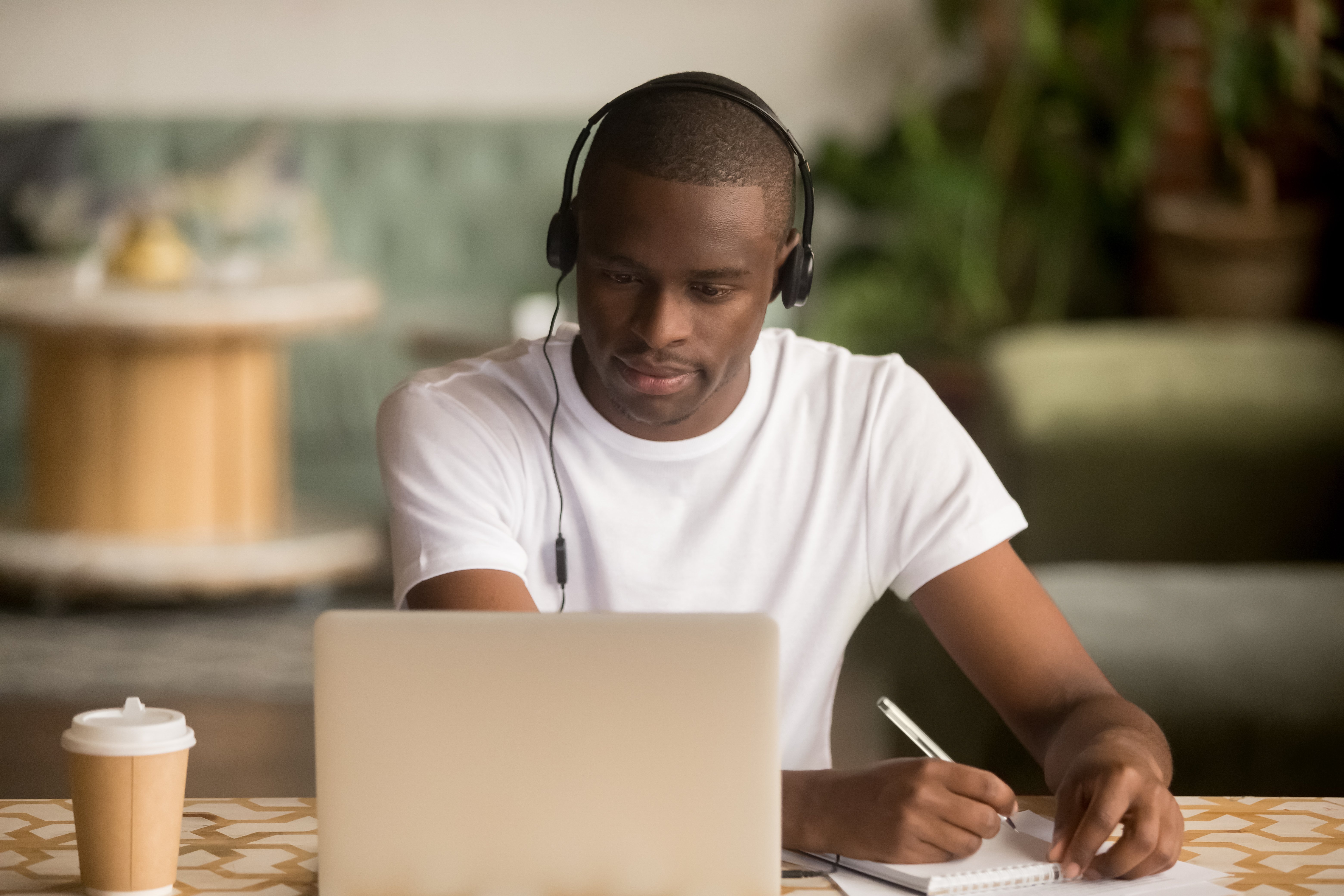 Male college student wearing headphones and making notes while watching laptop