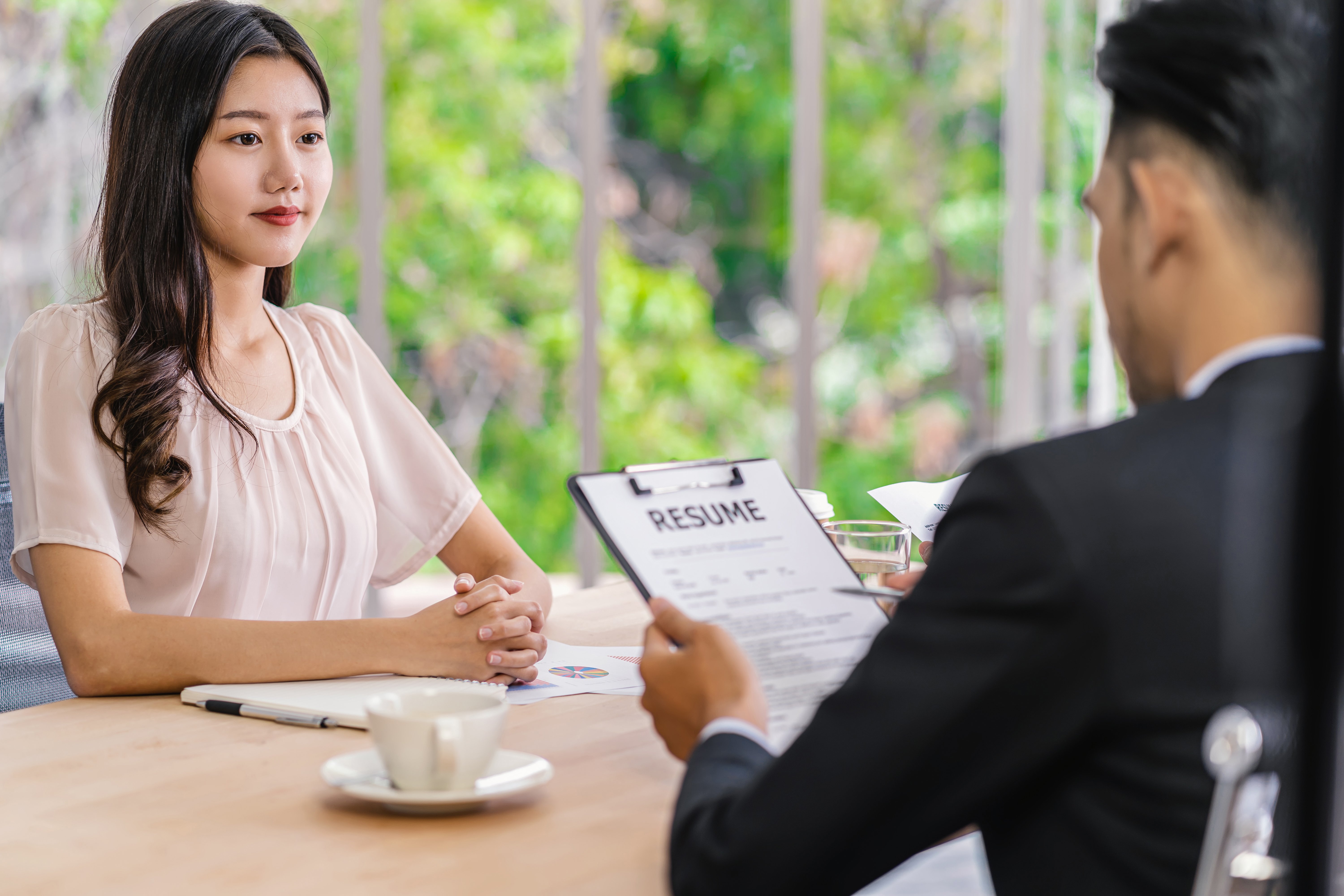 Woman with hands clasped sitting across a table from hiring manager holding clipboard