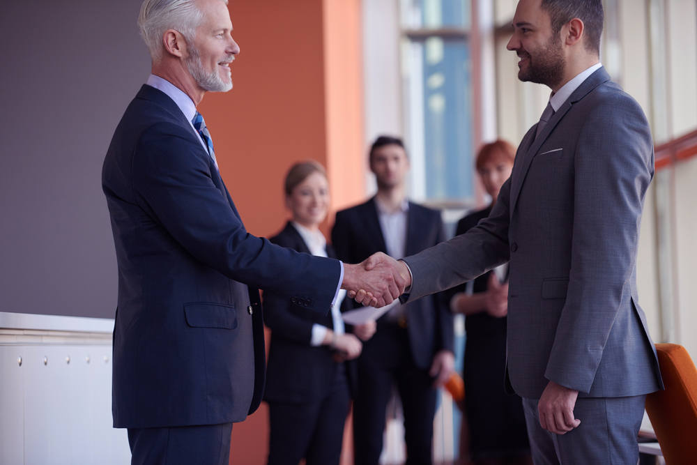 Two professionals shaking hands with a group of people in the background