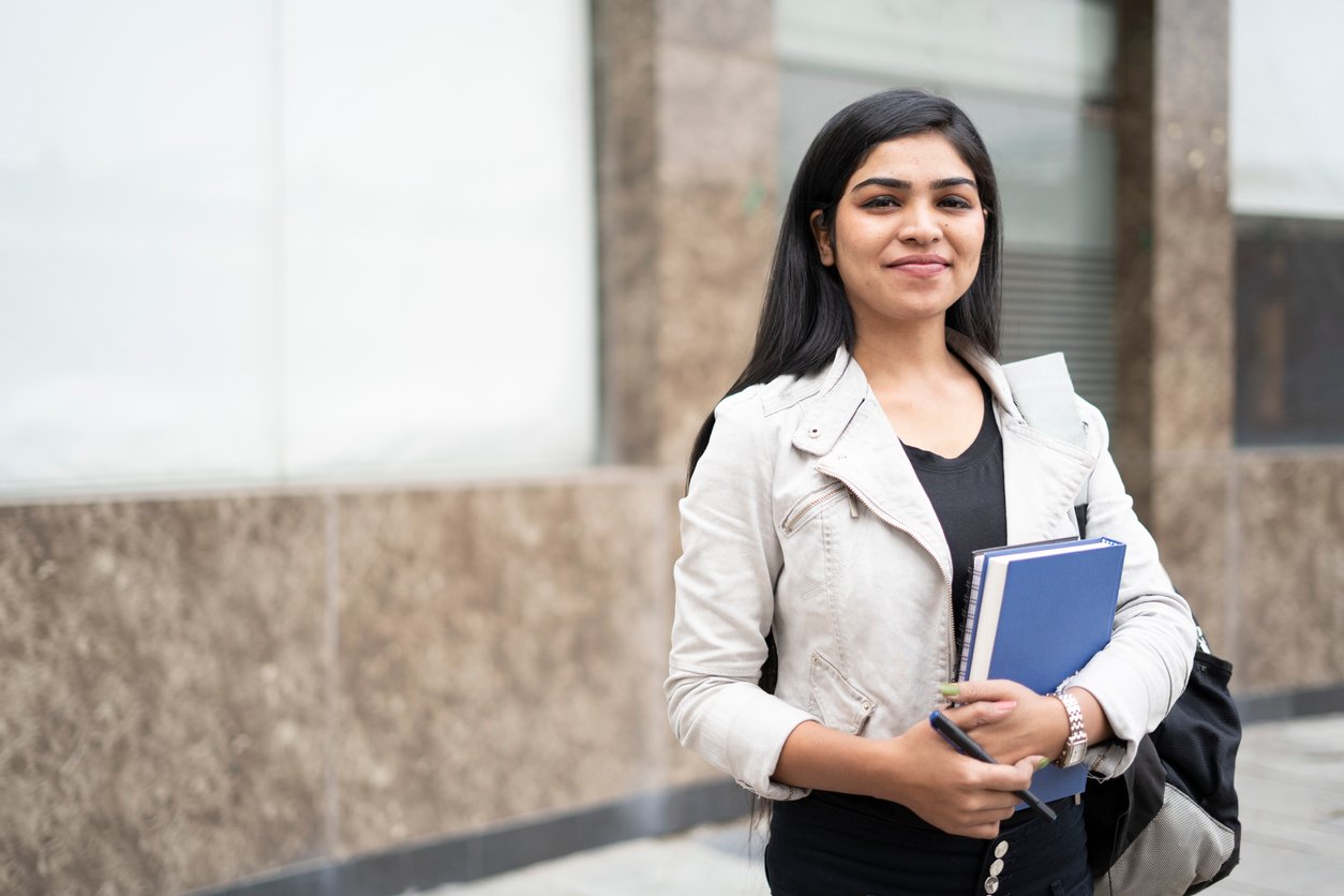 smiling woman carrying books and a backpack standing outside a building