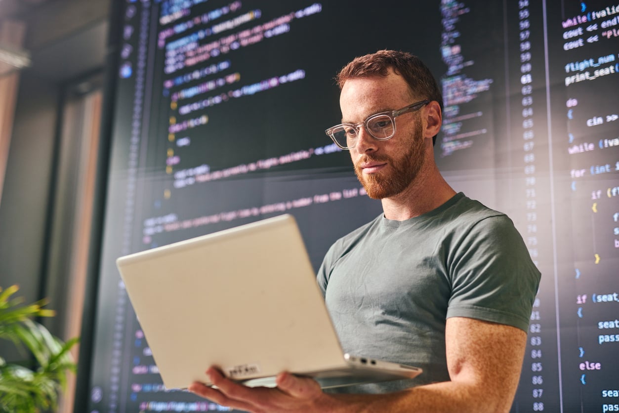Cybersecurity analyst working on laptop with code displayed on screen behind him