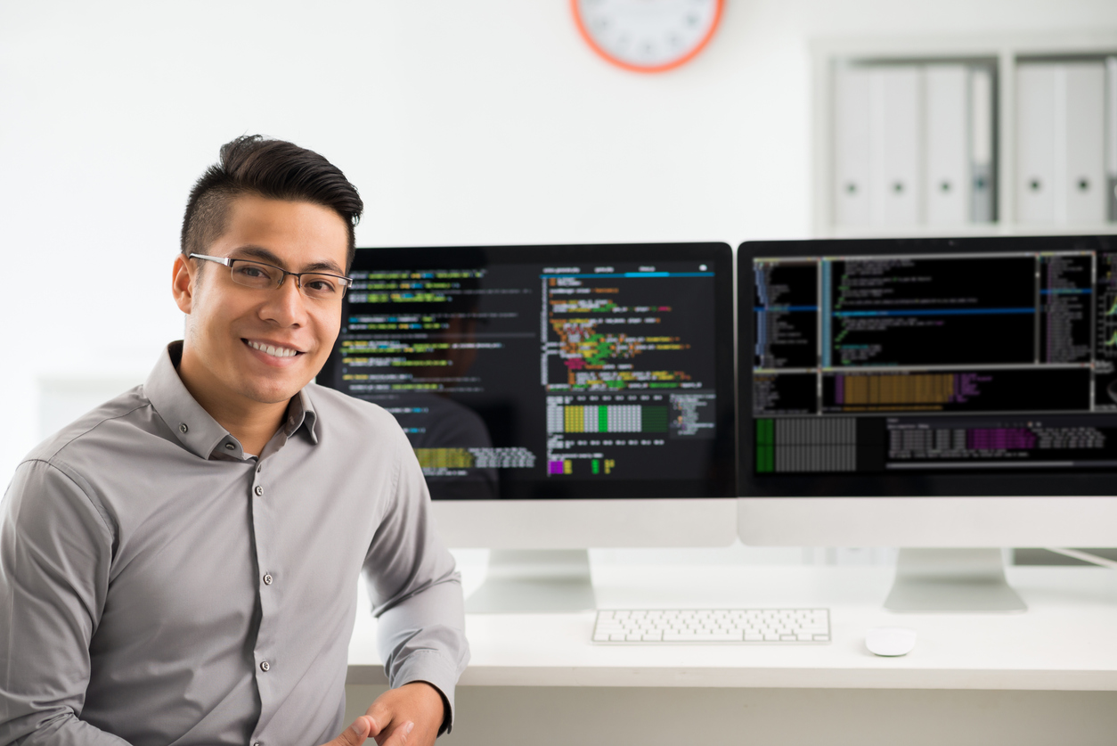 Smiling programmer sitting next to two computer screens