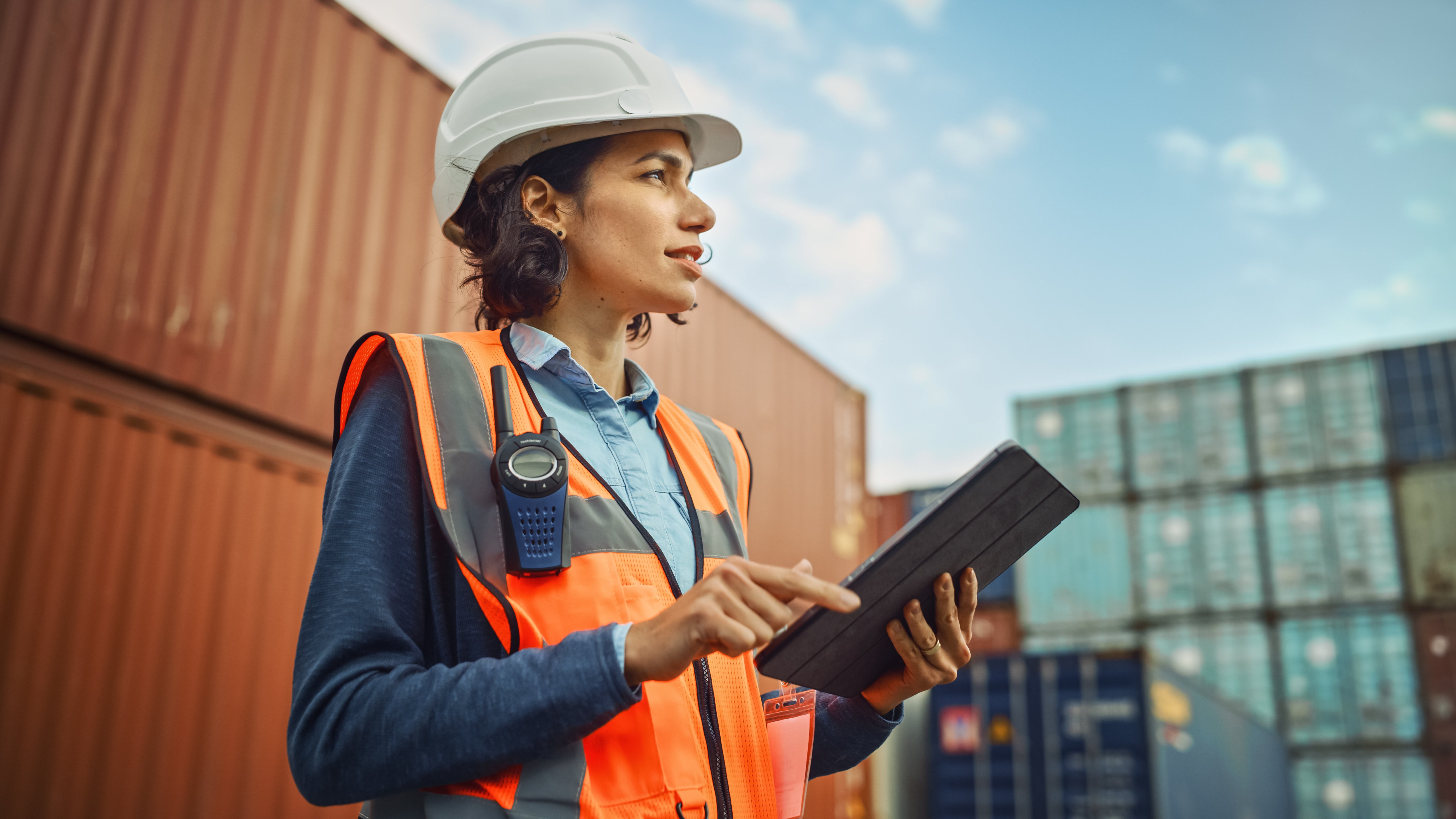 Woman in safety vest and hard hat standing near storage containers holding a tablet
