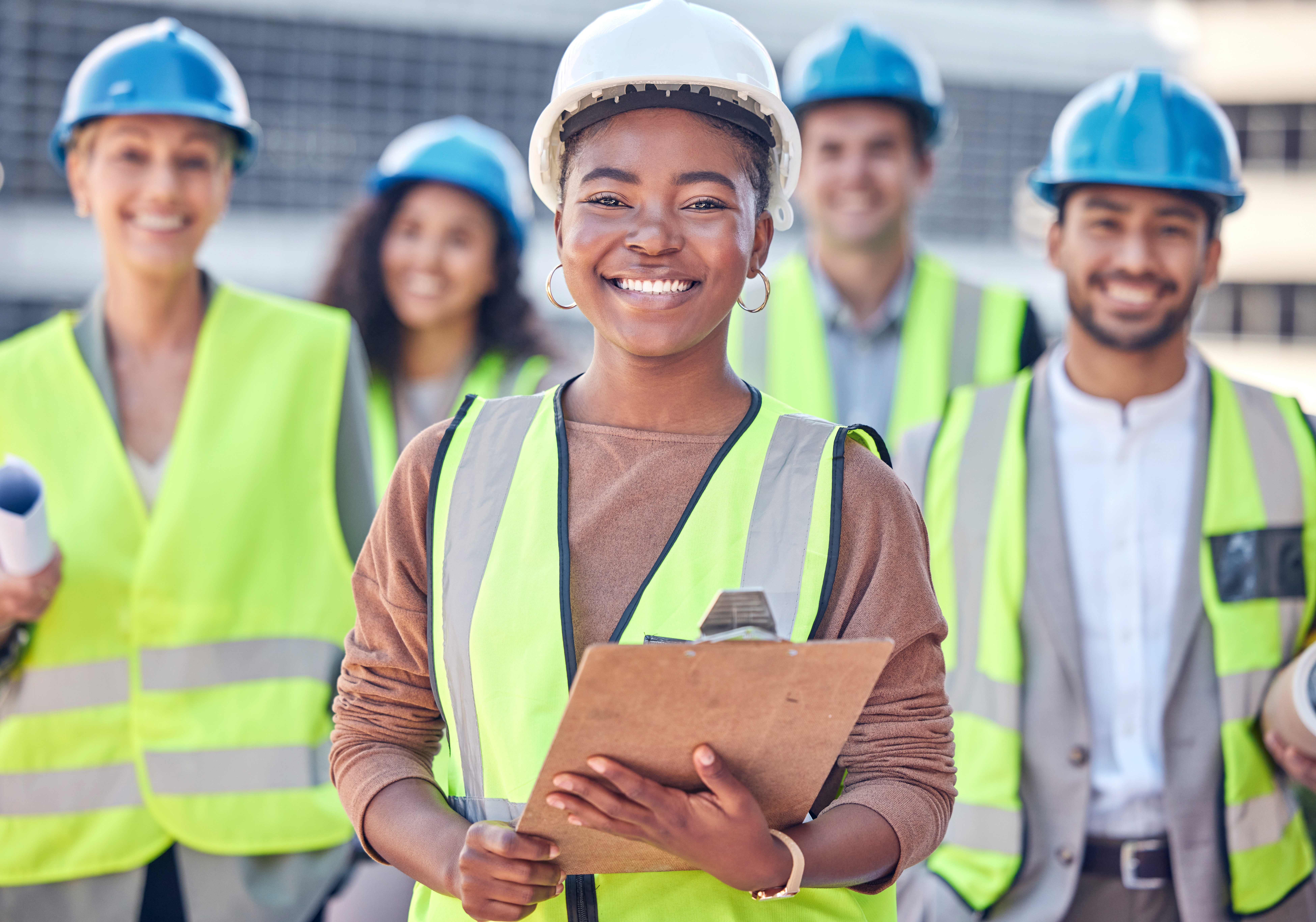 Construction worker holding clipboard and standing on building site with her colleagues in the background